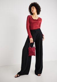 Rdeča majica z dolgimi rokavi in drapiranim izrezom, v paru z črnimi širokimi hlačami; drži majhno, teksturirano torbico s perlicami.