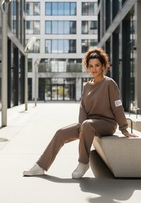 Brązowy dres z bluzą o okrągłym dekolcie i pasującymi spodniami. Biały tekst na rękawie, na tle nowoczesnego miejskiego krajobrazu. Białe trampki.