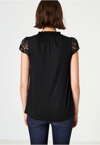 Černý top se shromážděným výstřihem a krátkými rukávy z krajky. Záda jsou z hladké látky, kombinované s tmavými džíny.