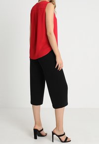 Punainen hihaton paita pyöreällä pääntiellä, yhdistetty mustiin mittatilaushyviin housuihin ja mustiin korkokengällisiin sandaaleihin. Sileä kangas, rento istuvuus.