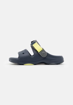 Granatowa sandałka na wsuwkę z dwoma szerokimi wycięciami i regulowanym żółtym paskiem na podbiciu, przedstawiona na białym tle.