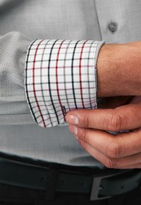 Camisa de cuadros gris con un puño de cuadros en blanco y colores. El puño presenta líneas rojas y azul marino. Mano ajustando la manga.