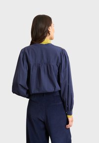 Blusa blu navy, con vestibilità ampia, maniche a palloncino e colletto verde a contrasto. Presenta una schiena arricciata e bottoni decorativi sulle maniche.