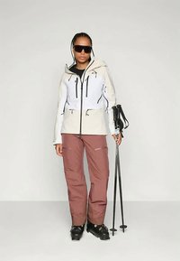 Femme portant des lunettes de soleil, une veste blanche et beige, un pantalon de ski marron, des chaussures de ski, tenant des bâtons de ski noirs, se tenant devant un fond uni.