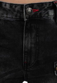 Black denim jeans with a metal button closure, red stitching accents, and a textured finish. Features standard pockets and a slim fit.