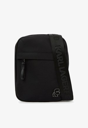 Čierna crossbody kabelka s predným vreckom so zipsom, hrubým popruhom s plastickým nápisom „KARL LAGERFELD“ a kovovým detailom loga v dolnom rohu.