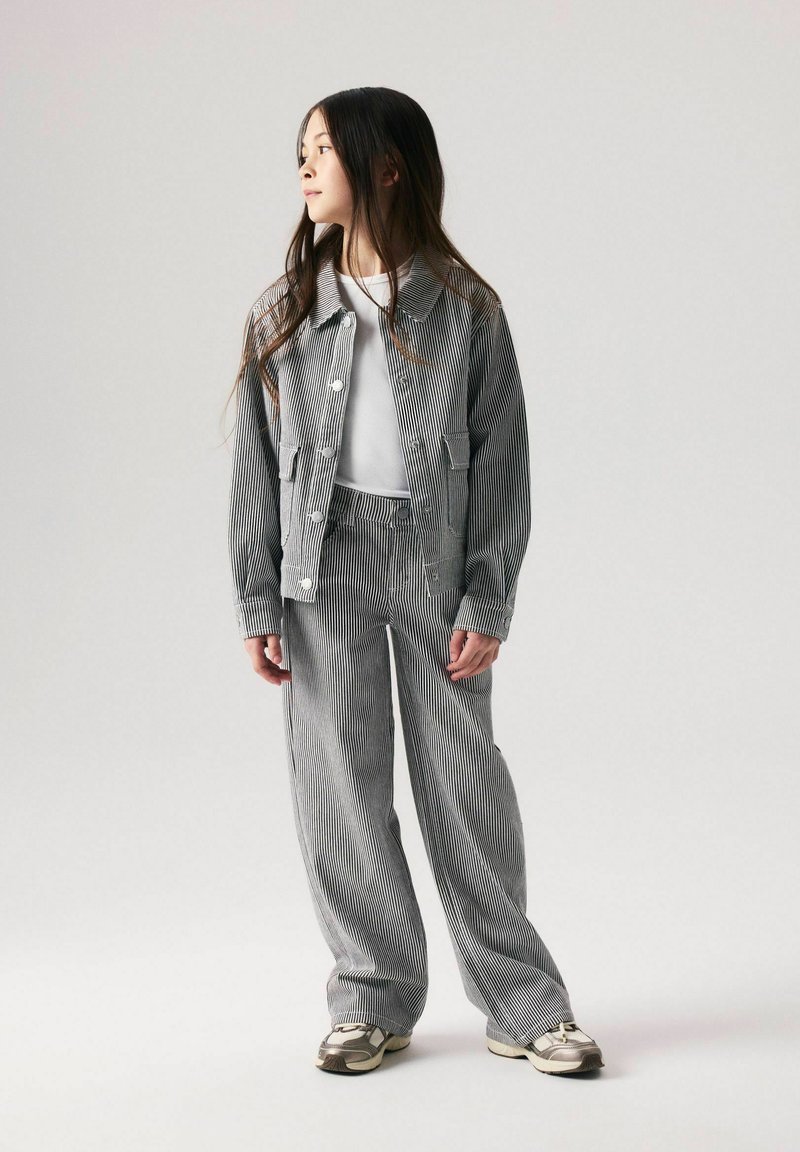 Jeune fille debout regardant vers la gauche, portant une veste grise et blanche à fines rayures et un pantalon large assorti avec des baskets blanches.