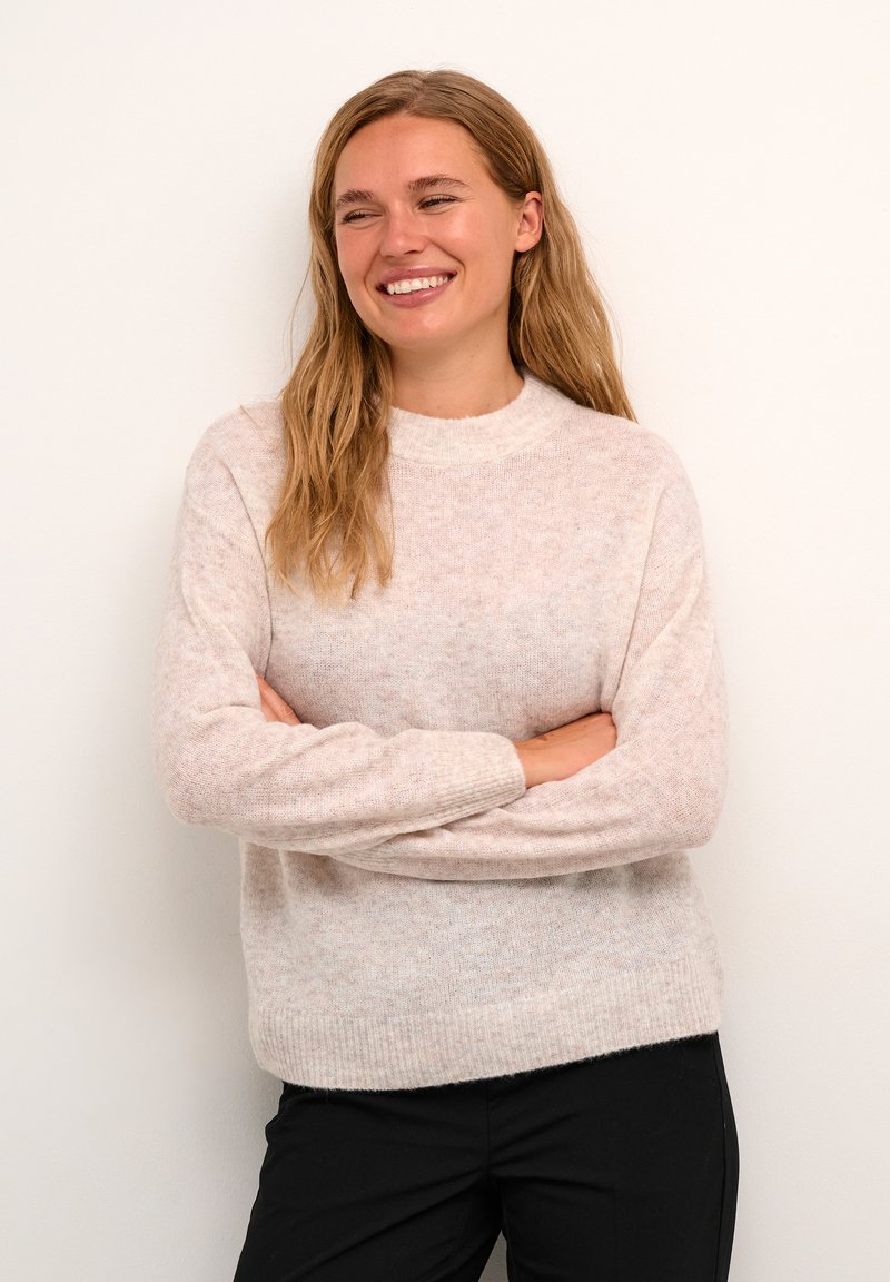 Helles beige gestricktes Sweatshirt mit rundem Halsausschnitt, langen Ärmeln und gerippten Bündchen. Weiche Textur und leicht lockere Passform. Über einer schwarzen Hose getragen.