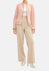 Jasnoróżowa kurtka zapinana na zamek, wykonana z miękkiego materiału, z okrągłym dekoltem i dwoma bocznymi kieszeniami, zestawiona z beżowymi spodniami o szerokich nogawkach oraz białymi botkami na obcasie.
