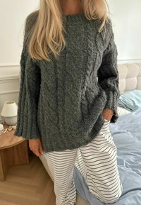 Maglione chunky verde scuro lavorato a maglia con un design a trecce e maniche larghe, abbinato a pantaloni a gamba larga bianchi a righe.