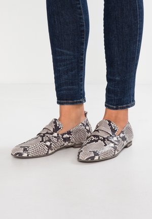 Osoba w niebieskich dżinsach i szarych loafersach w wzór skórki węża, stojąca na gładkiej białej podłodze.