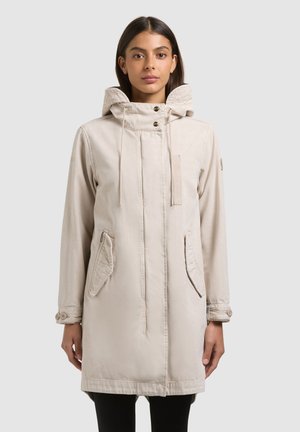 Femeie purtând o geacă parka bej cu glugă, buzunare frontale și manșete cu nasturi, stând în fața unui fundal simplu, gri deschis.