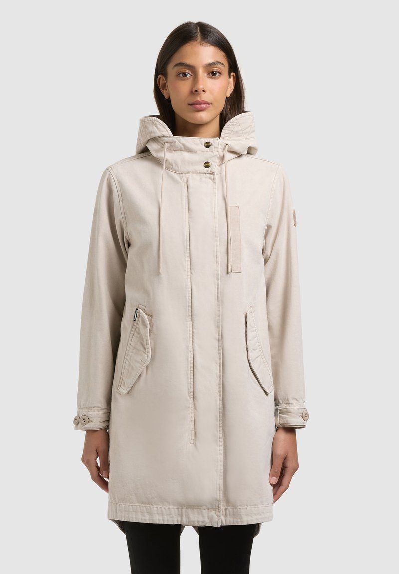 Mujer con abrigo parka beige con capucha, bolsillos delanteros y puños con botones, posando frente a un fondo liso de color gris claro.