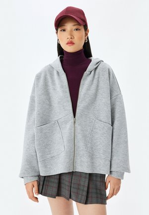 Sweat à capuche gris zippé avec une coupe oversize, poches devant et capuche. Porté par-dessus un col roulé bordeaux, associé à une jupe plissée à carreaux.