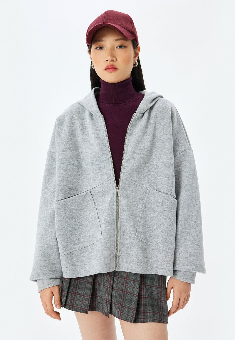 Grå zip-up hættetrøje med oversized pasform, forlommer og hætte. Båret over en burgunder turtleneck, parret med en ternet plisseret nederdel.