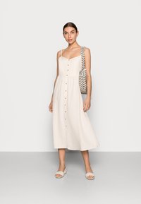 Beiges Leinen-Midikleid mit Spaghettiträgern, Knöpfen vorne und einem taillierten Schnitt. Kombiniert mit einer gemusterten Tasche und hellen Sandalen.