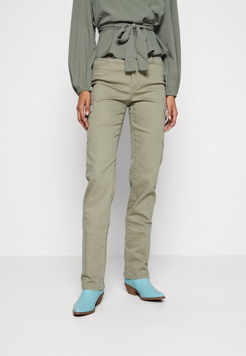 Femme portant un pantalon cargo vert olive et un haut assorti noué à la taille, associé à des bottines bleu clair à talons en bois.