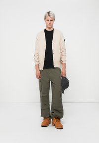 Beige cardigan over a black shirt, olive cargo pants, and tan boots, holding a gray cap. The cardigan features buttons and a patch.
