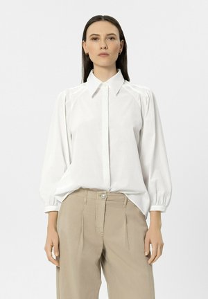 Frau mit glattem dunklem Haar, die eine weiße Bluse mit langen Ärmeln und gepufften Schultern sowie beige High-Waist-Hosen trägt und vor einem schlichten Hintergrund steht.