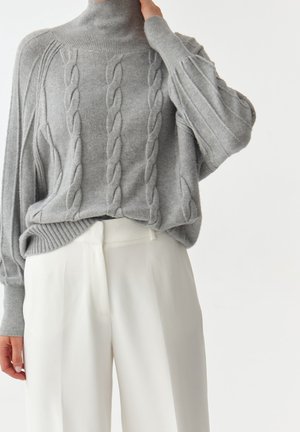 Pull gris à col roulé en maille torsadée avec manches bouffantes, associé à un pantalon blanc taille haute ample porté par une personne debout.