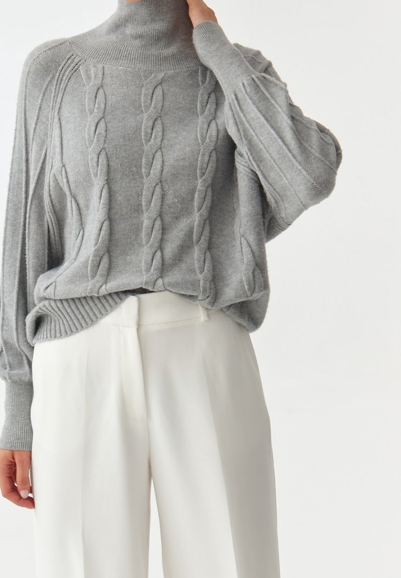 Pull gris à col roulé en maille torsadée avec manches bouffantes, associé à un pantalon blanc taille haute ample porté par une personne debout.