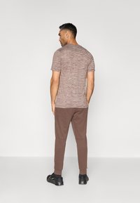 Mand, der vender væk, iført en kortermet meleret brun t-shirt, brune joggerbukser, sorte sokker og sorte sportssko.