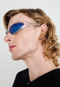 Occhiali da sole trasparenti con un design sportivo, caratterizzati da montature chiare e lenti con tinta blu. Marchio ben visibile sui lati, finitura liscia.