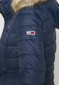 Tmavě modrá prošívaná péřová bunda s kapucí lemovanou kožešinou, zdobená horizontálním prošíváním a bílou, červenou a modrou nášivkou na rukávu.