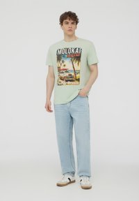 Maglietta verde chiaro con un design retro di surf stampato. Indossata con jeans azzurri chiari e sneakers bianche con accenti neri e marroni.