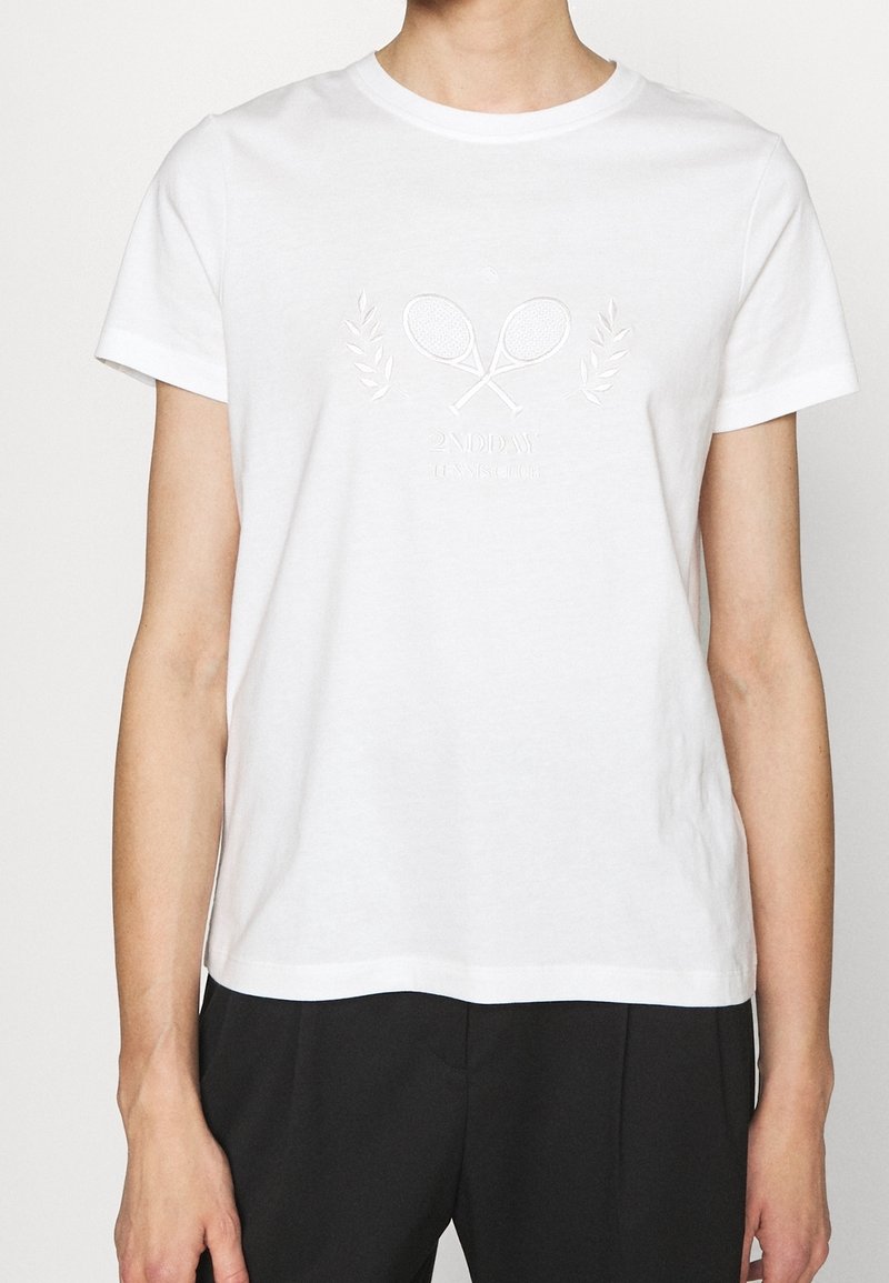 Personne portant un t-shirt blanc avec un design en relief de raquettes de tennis croisées et de lauriers, assorti à un pantalon noir, coupé aux épaules et à la taille.