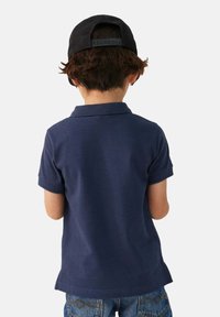Donkerblauwe polo met een kraag, korte mouwen en een afgeronde zoom, gecombineerd met blauwe spijkerbroek. Achteraanzicht toont de gestructureerde stof.