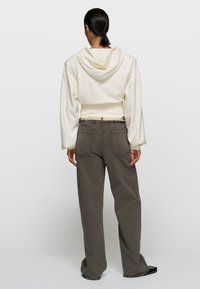 Frau mit zurückgebundenem Haar, die einen cremefarbenen, kürzeren Hoodie und lockere graue Hose trägt und vor einem einfarbigen Hintergrund mit dem Rücken zur Kamera steht.