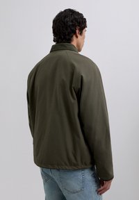 Man standing with back turned wearing an olive green jacket and light blue jeans against a plain gray background.