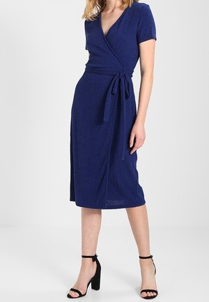 Woman wearing a knee-length, short-sleeve blue wrap dress with a tied waist and black high-heeled ankle-strap sandals.