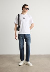 T-shirt blanche avec un logo noir, jeans bleus, baskets blanches et lunettes de soleil noires. Le sac est porté en diagonale sur le corps.