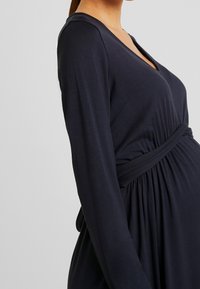 Marinblå klänning med långa ärmar och V-ringning samt rynkad midjedetalj, tillverkad av mjukt, stretchigt material som rymmer en växande figur.