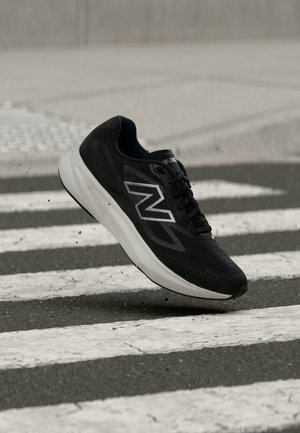 Chaussure de course New Balance noire et blanche en l'air au-dessus d'un passage piéton sur une route asphaltée.