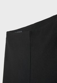 Gros plan de la ceinture et de la partie supérieure d’un vêtement en tissu noir uni, probablement une jupe ou un pantalon, sur un fond clair uni.