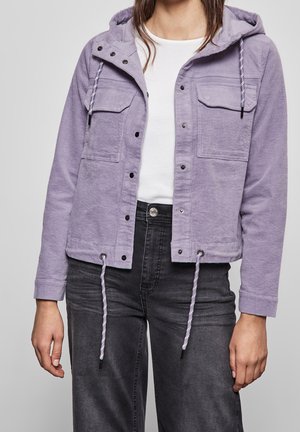 Personne portant une veste en velours côtelé violet clair avec poches avant et cordons, chemise blanche et jean gris foncé sur un fond neutre.