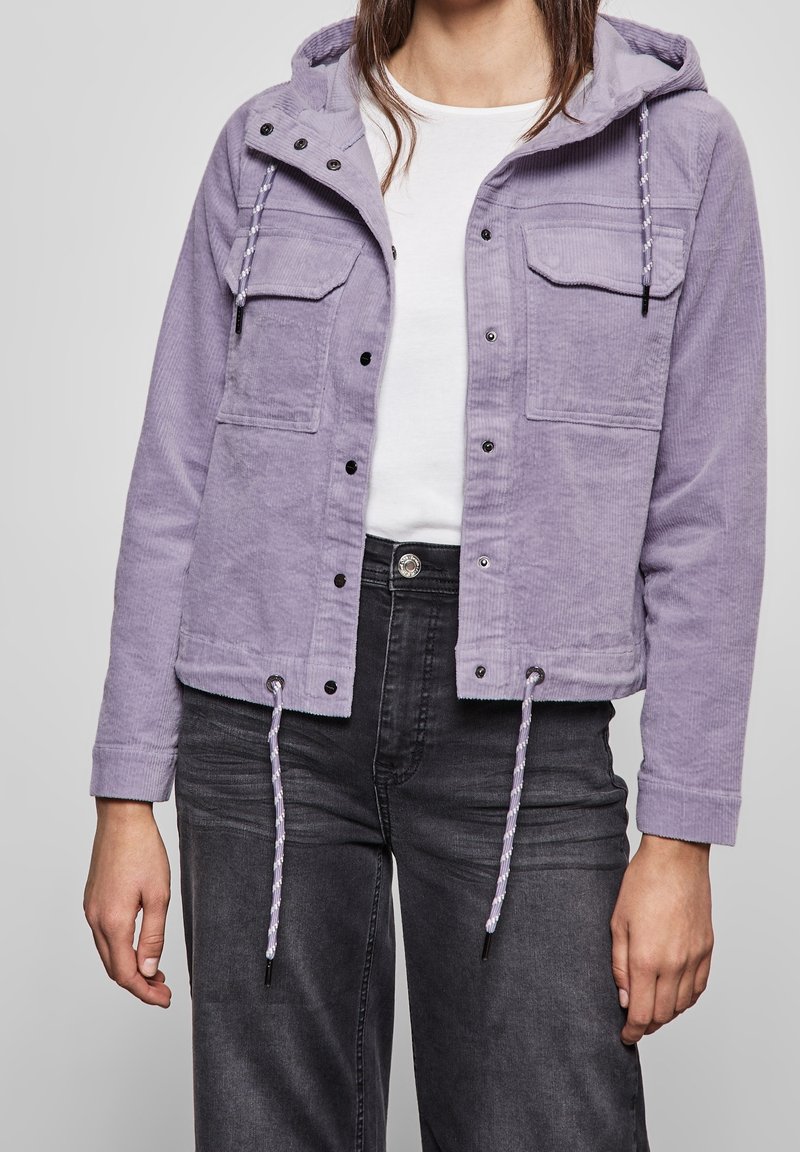 Personne portant une veste en velours côtelé violet clair avec poches avant et cordons, chemise blanche et jean gris foncé sur un fond neutre.