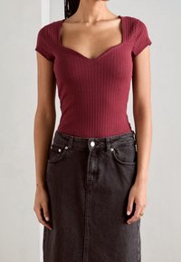 Donna che indossa un top corto a manica corta, aderente e a coste, di colore bordeaux con scollatura quadrata, abbinato a una gonna di jeans nera a vita alta.