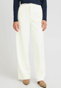 Pantalons larges blancs avec une texture lisse, dotés d'une fermeture à bouton et de poches. Associés à un haut à manches longues bleu marine et des chaussures beiges.