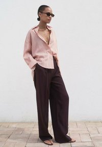 Chemise oversize rayée rose clair avec des manches larges, associée à un pantalon sombre ample et des sandales plates, sur fond de mur blanc uni.