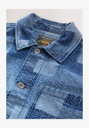 Camicia in denim patchwork blu con cuciture strutturate, bottoni in metallo, una tasca sul petto e un'etichetta con la scritta "Premium Grade 1982 Next".