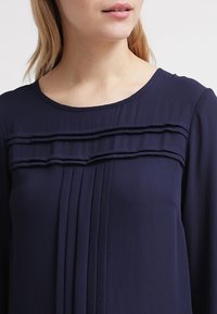 Blusa blu navy con scollatura rotonda, caratterizzata da ruffles sovrapposti sul petto e pieghe verticali per un tocco di texture e design.