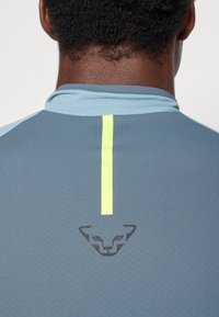Cămașă sport de culoare albastru deschis, cu o fâșie verticală galbenă contrastantă și un logo închis la spate, confecționată dintr-un material texturat.