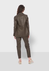 Femme debout de dos, portant un blazer en cuir marron foncé et un pantalon assorti, avec des baskets claires sur un fond uni.