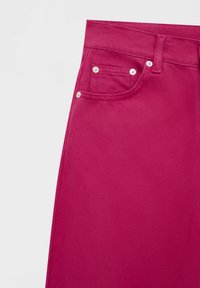 Tissu en denim rose vif montrant une poche avant, une poche à monnaie, un passant de ceinture et des rivets argentés sur un fond uni clair.