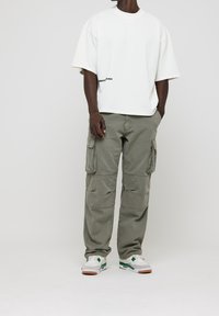 Camiseta blanca de gran tamaño con gráfico negro, pantalones cargo verde oliva con bolsillos laterales y zapatillas blancas con acentos en verde y gris.