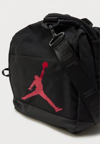 Μαύρη τσάντα από ανθεκτικό υλικό, με το χαρακτηριστικό κόκκινο λογότυπο Jumpman. Διαθέτει φερμουάρ στην κορυφή και ρυθμιζόμενο λουρί με επωνυμία.